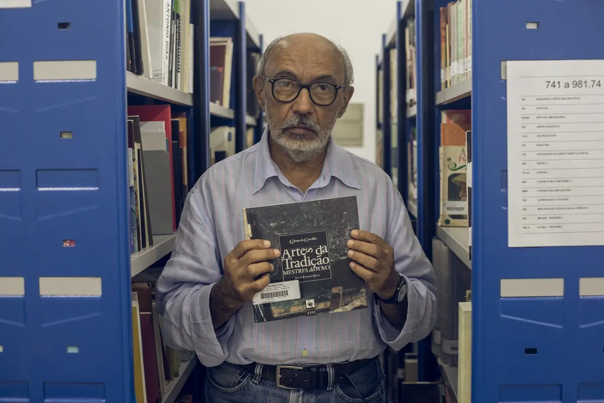 Gilmar de Carvalho segurando livro entre estantes