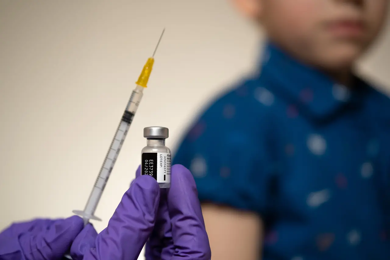 Mão com luva segura dose de vacina e injeção, enquanto criança aparece atrás, embaçada
