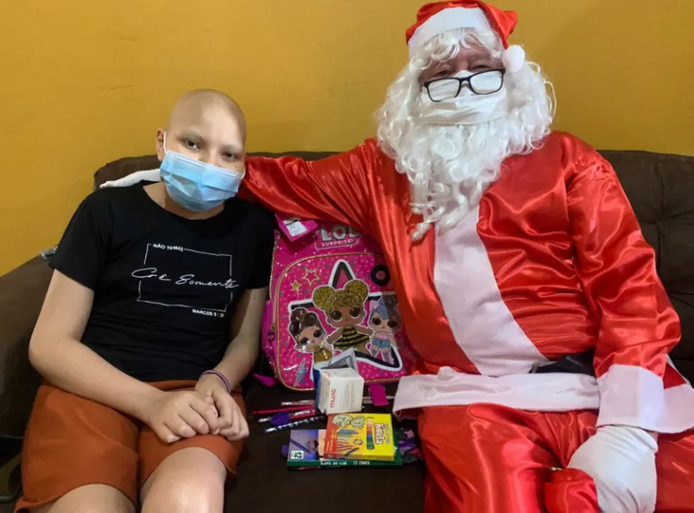 Tailane Alves da Silva sentada em um sofá ao lado do Papai Noel e de seu presente