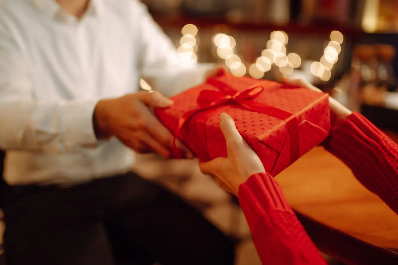 Mãos tocando uma caixa de presente vermelha, com luzes de Natal ao fundo