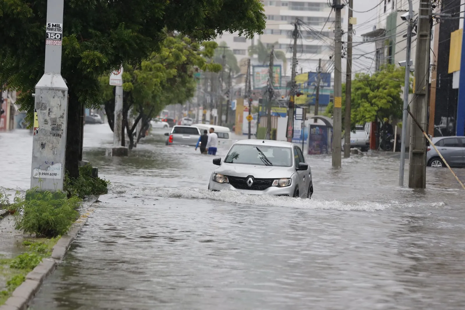 Carros, pedestres e motociclistas passam por avenida alagada na Capital