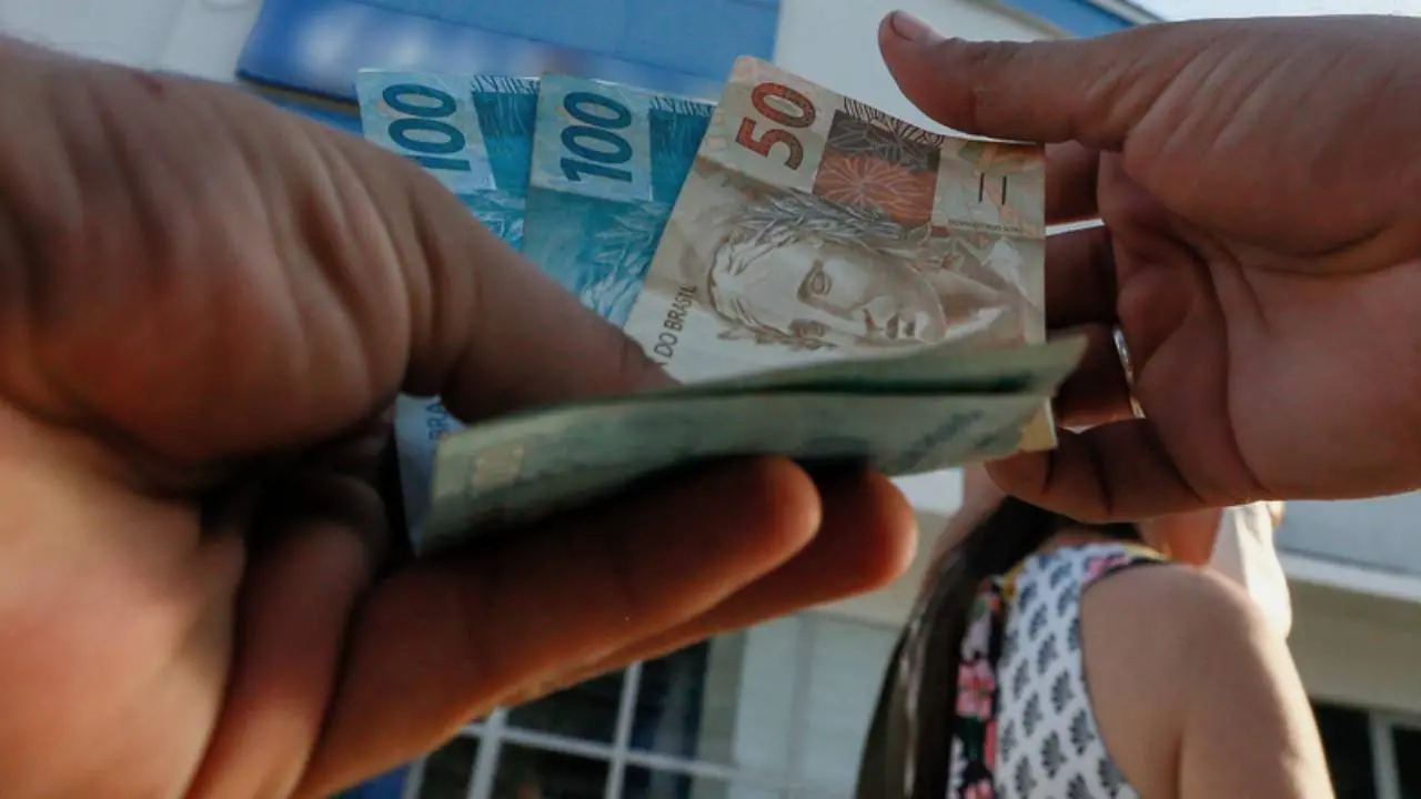 Mãos segurando notas de real em frente a agência bancária