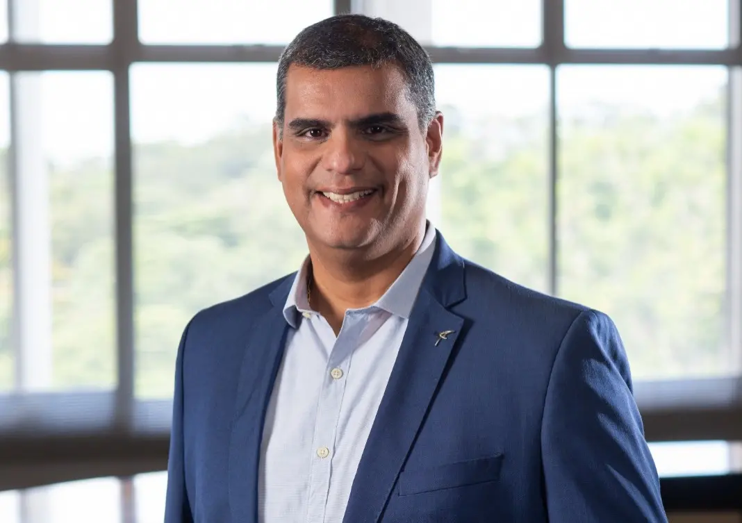 Erick Torres é um homem negro de pele clara, alto, de cabelos curtos e grisalhos. Na foto, ele está sorrindo e usa um terno azul por cima de uma camisa social da mesma cor