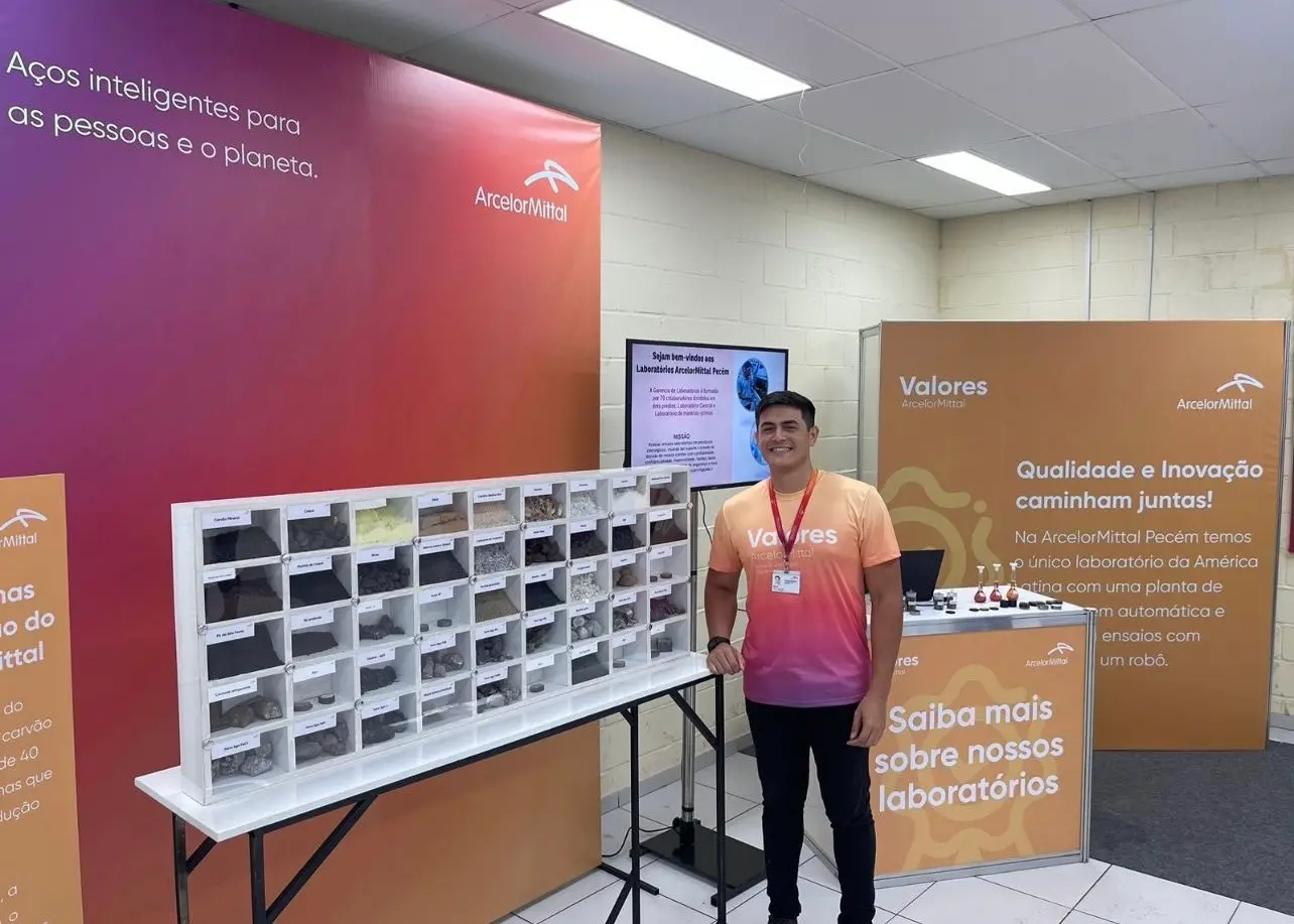 Alex é um homem branco, alto, de cabelo preto. Na foto, ele está posando ao lado de materiais para a produção do aço