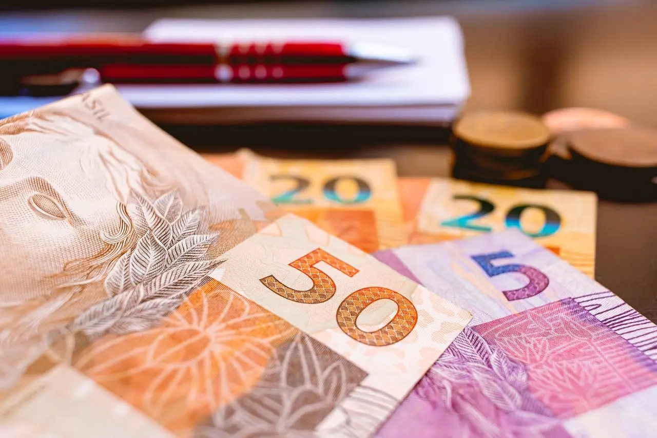 Notas e moedas de Real sobre uma mesa com um caderno e uma caneta