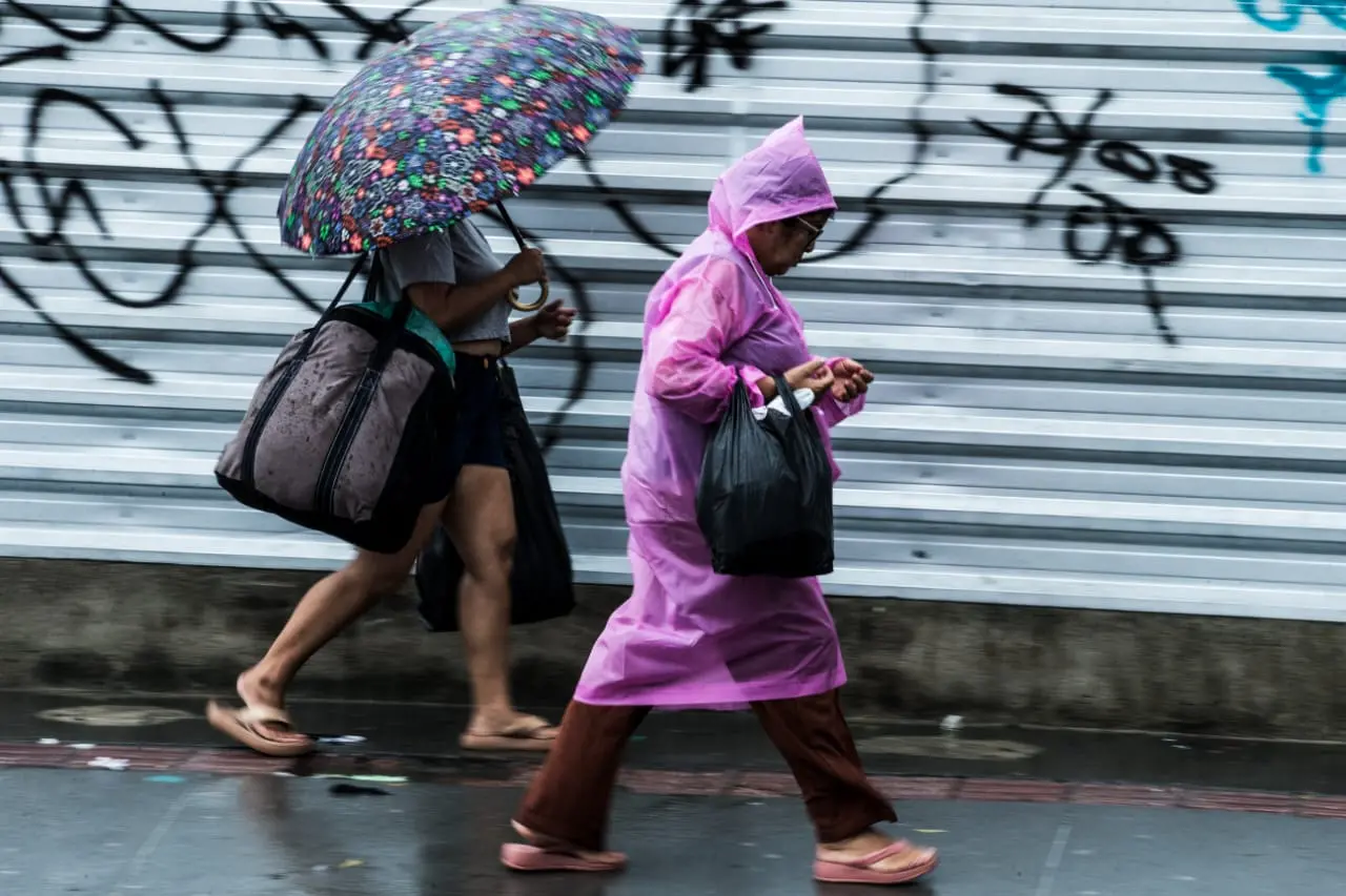 Duas pessoas andando na rua sob chuva de Fortaleza, com uma usando capa de chuva rosa e a outra segurando um guarda-chuva colorido. Uma imagem que retrata o cotidiano em dias chuvosos nas cidades.