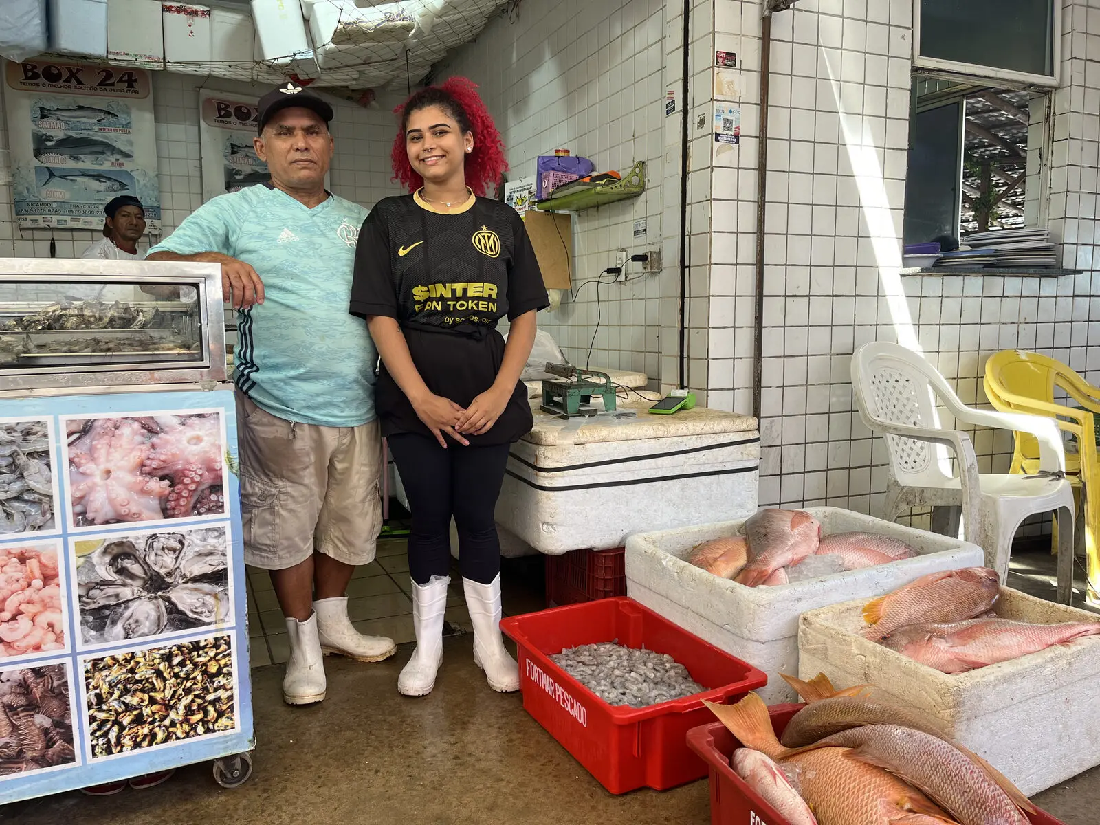 Yorrana ao lado do pai, com caixas de peixes e camarões à frente deles, em um box no Mercado dos Peixes de Fortaleza