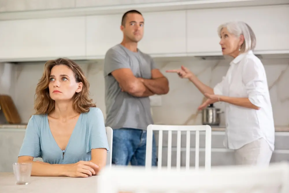 Mulher sentada na mesa da cozinha triste enquanto marido e sogra discutem no plano secundário