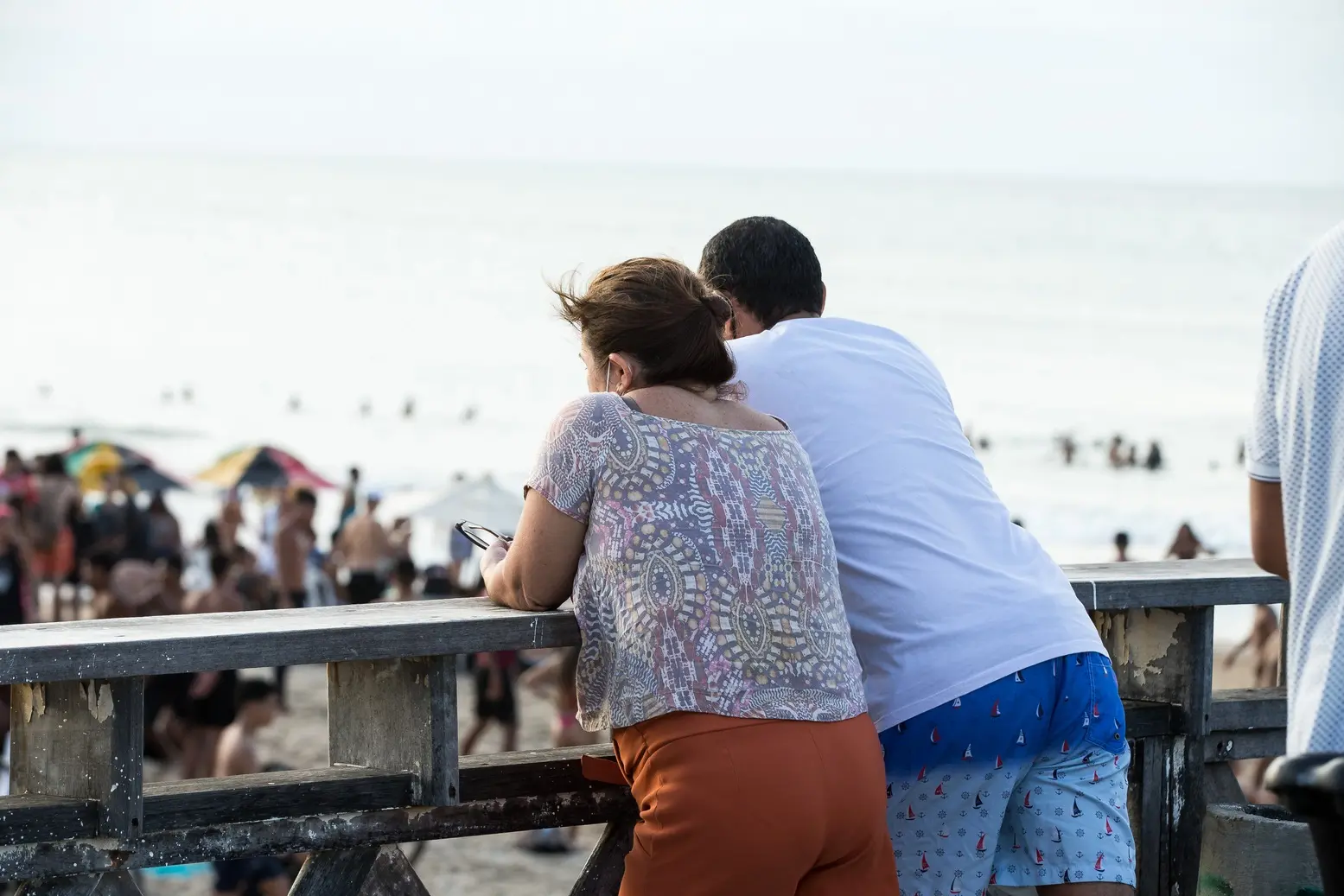 Casal apoiado em vigas de um espigão em Fortaleza numa tarde com pessoas ao fundo na areia da praia e no mar