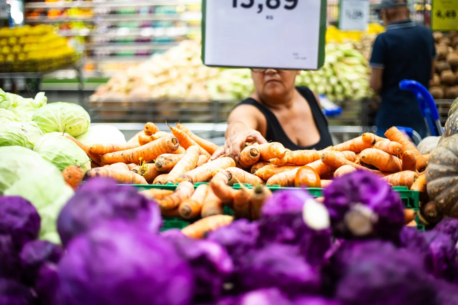 Imagem mostra cenoura e repolho expostos em um supermercado. Uma mulher fazendo compras tenta alcançar uma das cenouras expostas