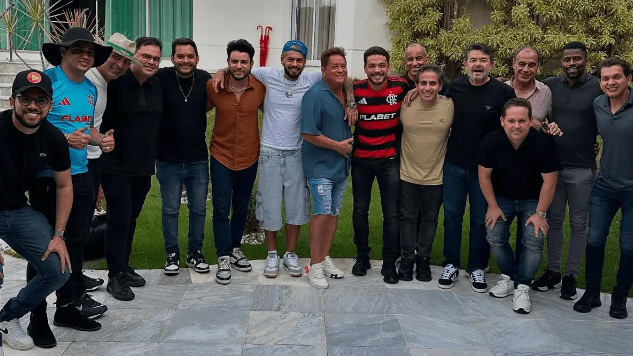 Grupo de dezesseis homens posando sorridentes para uma foto em área externa, em frente a um prédio com janelas e plantas. Entre eles estão os cantores Wesley Safadão e Leonardo, que aparecem no centro da imagem com roupas casuais. Wesley usa camiseta branca e boné azul, enquanto Leonardo veste camisa azul e bermuda jeans. Outro homem ao lado usa camisa do Flamengo. A maioria dos presentes está de calça jeans e camiseta, alguns com chapéu. A atmosfera é descontraída e informal