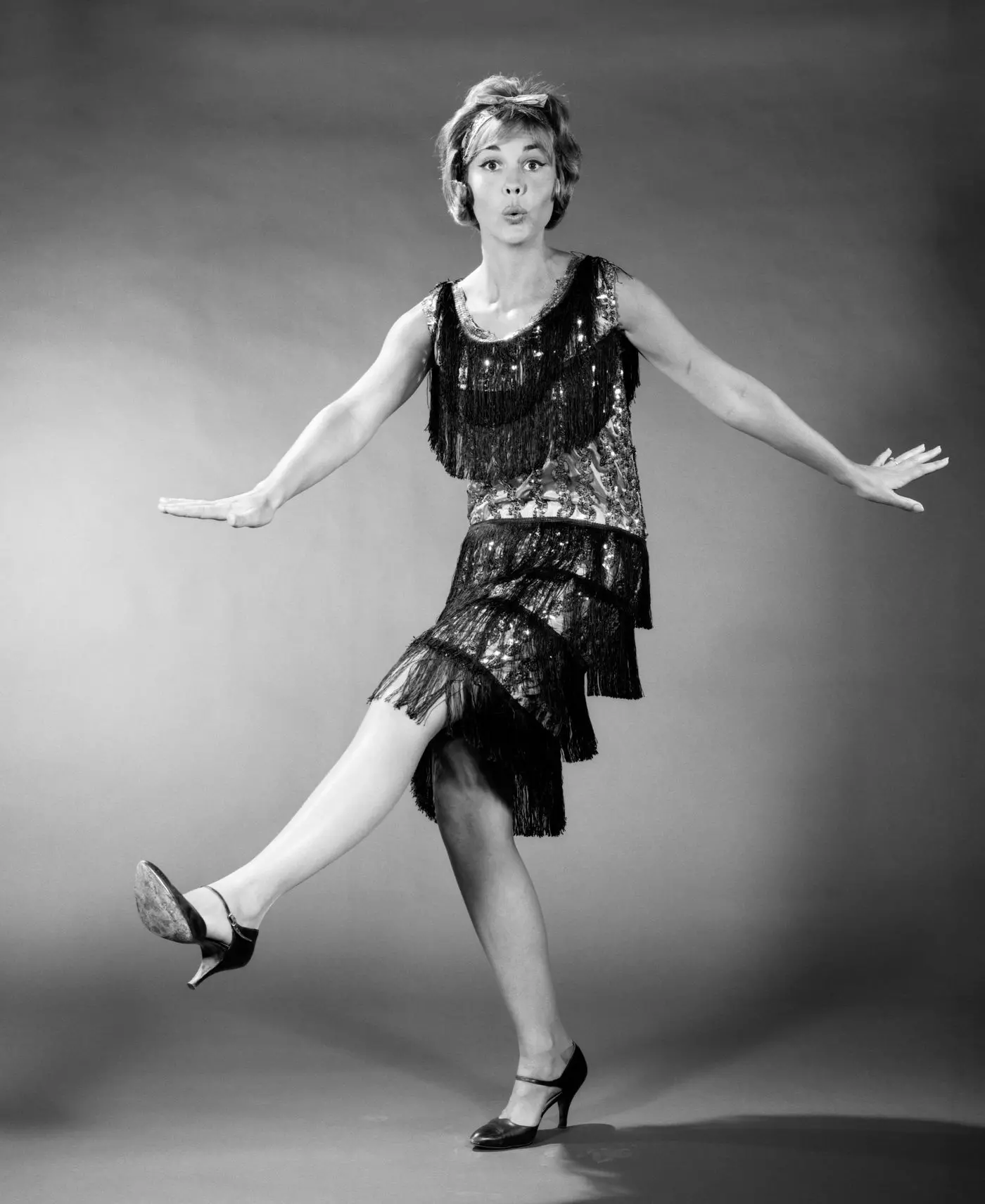 Foto histórica em preto e branco representando melindrosas dos anos 1920. Moça com cabelos presos em coque estilo chanel, vestido de franjas e oças de saias curtas