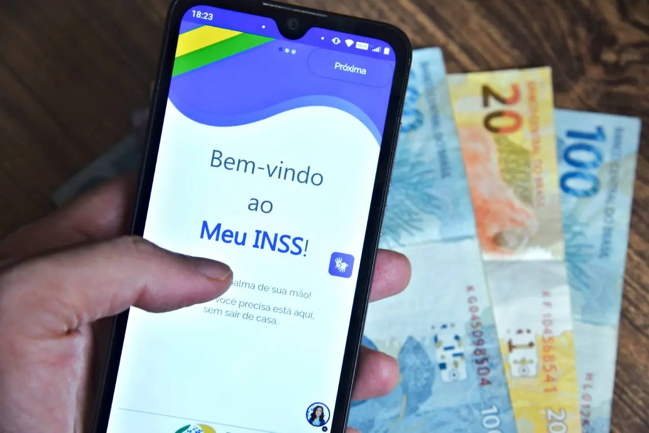 Mão segurando celular com aplicativo Meu INSS e notas de Real ao fundo no contexto da fraude investigada