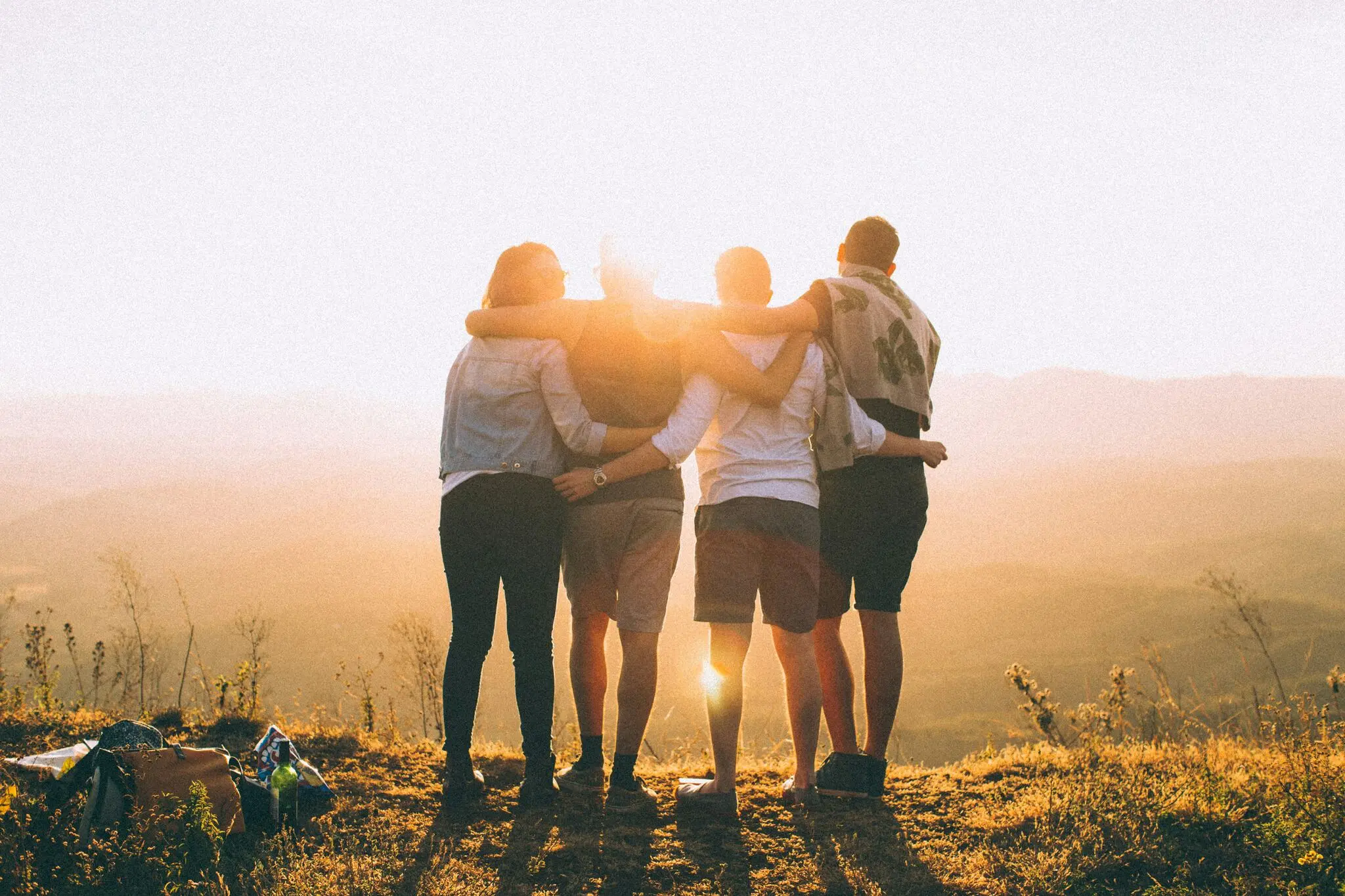 Grupo de quatro pessoas de costas para a câmera, se abraçando e contemplando a vista de um local alto, na natureza, ao pôr-do-sol