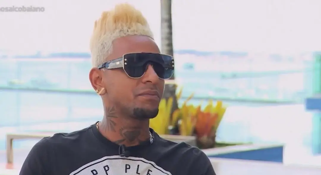 Imagem de um homem com cabelo loiro platinado e óculos escuros, posando em ambiente moderno e iluminado, representando estilo e atitude.