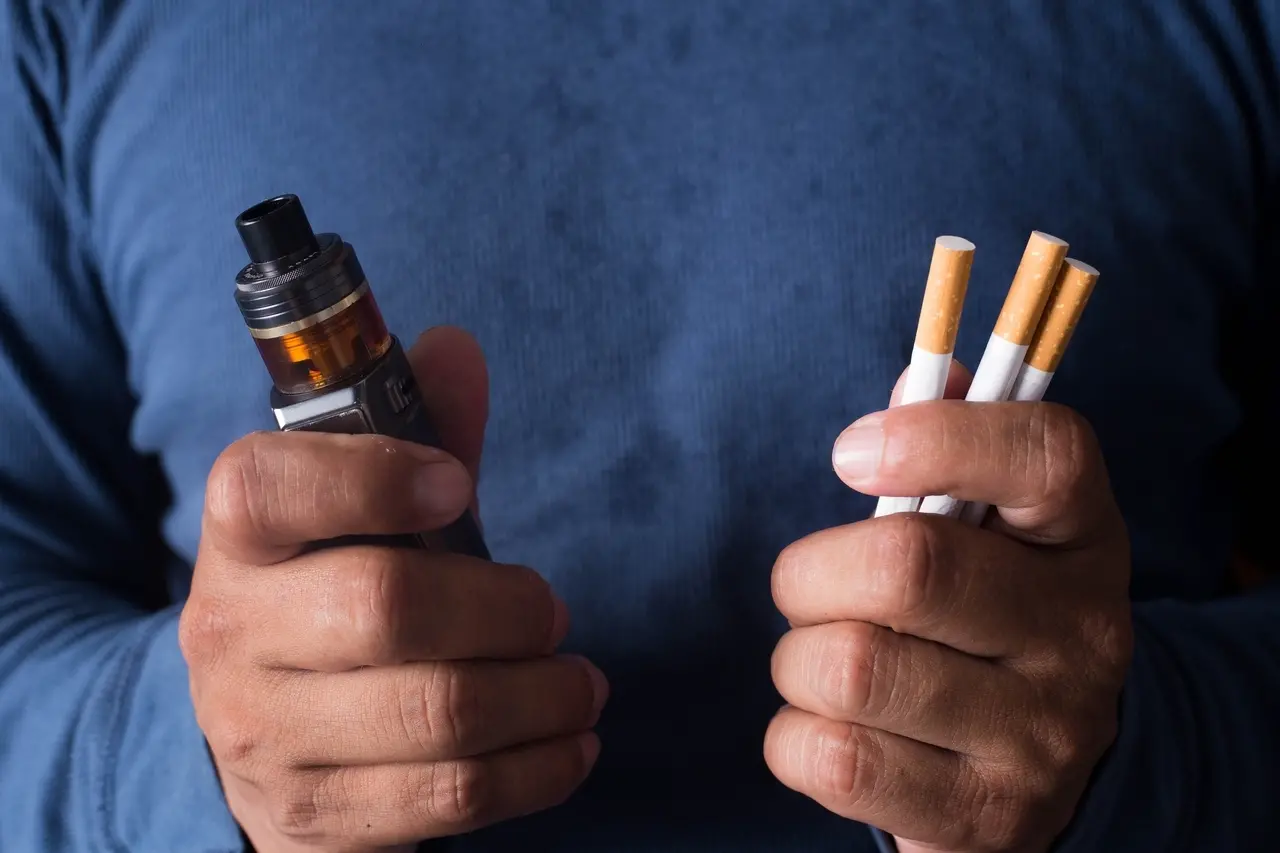 Imagem mostra homem jovem segurando cigarro eletrônico (ou vape, ou pod) e cigarro tradicional, sobre fundo escuro, ilustrando como parar de fumar em dia mundial sem tabaco