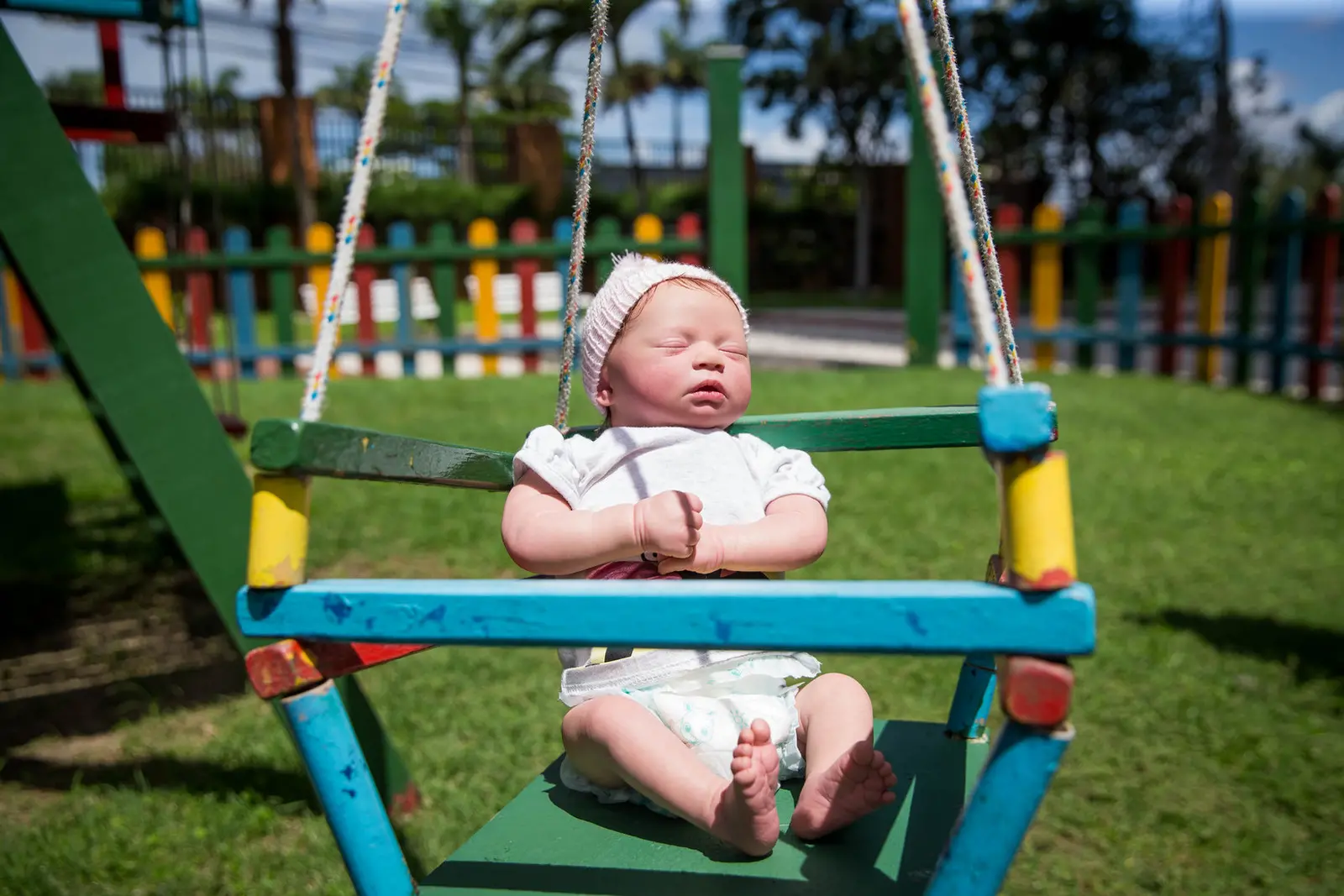 A imagem mostra bebê reborn sentado em um balanço colorido de madeira em um parque. O boneco veste um macacão branco, uma fralda e um gorro de tricô claro. Seus olhos estão fechados e as mãos estão juntas na frente do corpo. Ao fundo, há um gramado verde, uma cerca colorida de madeira e árvores, com estruturas de playground visíveis. O sol está batendo no rosto do boneco.