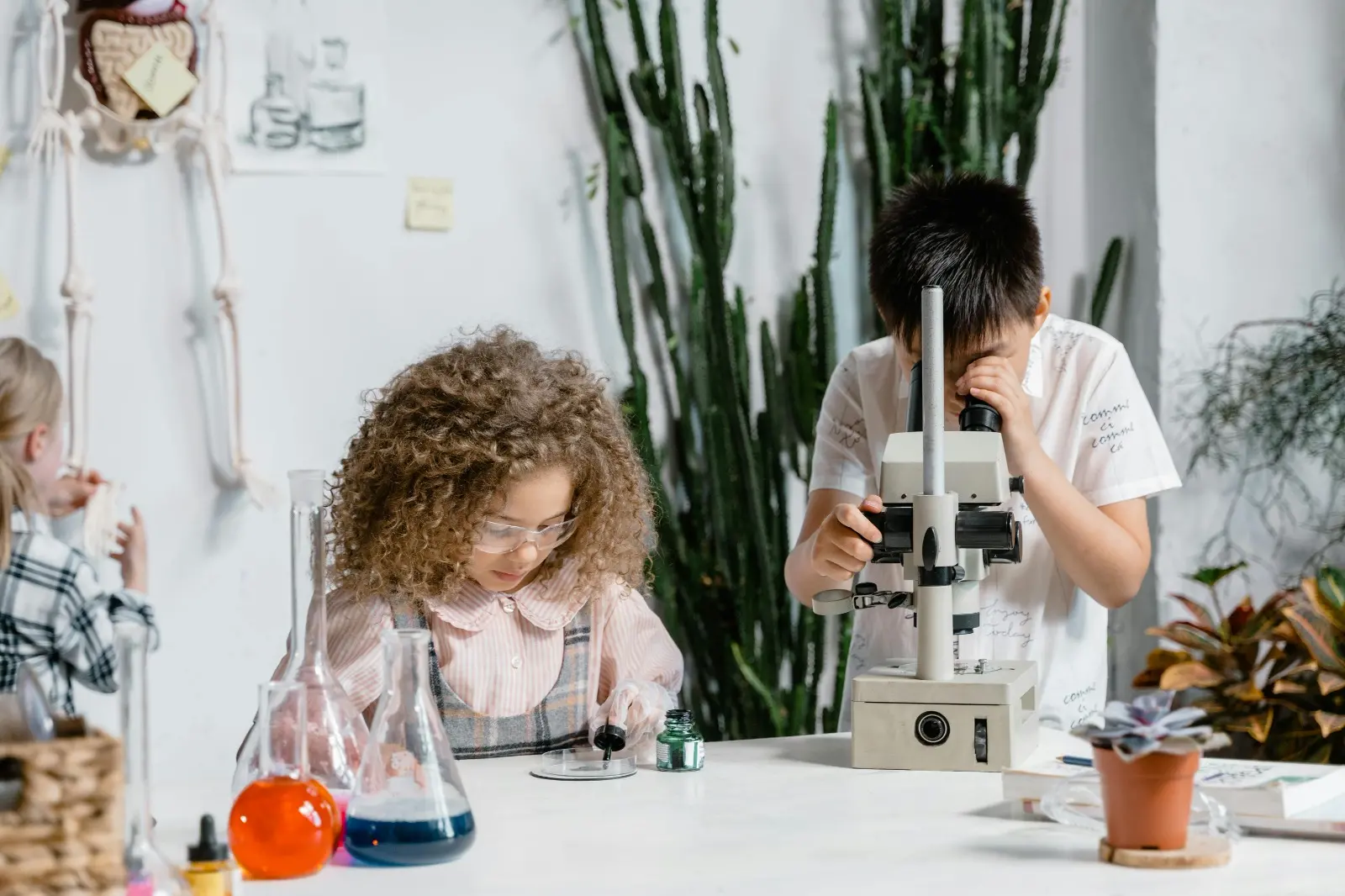 Duas crianças, uma menina de um menino, exploram experimentos em sala de ciências, com microscópio e frascos coloridos sobre a mesa.