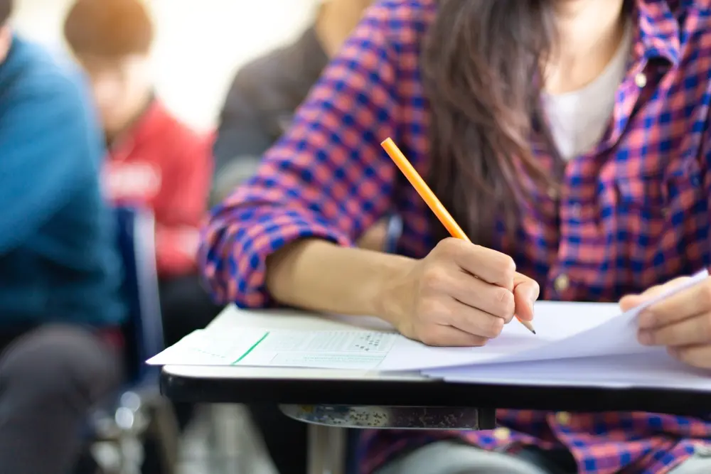 Estudante fazendo inscrição ou preenchendo formulários em uma sala de aula, foco na mão com caneta em papel, ambiente escolar ao fundo.