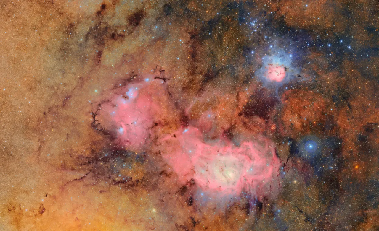 Imagem colorida do espaço profundo, mostrando três nebulosas brilhantes. O fundo é repleto de estrelas e nuvens de poeira em tons dourados, marrons e alaranjados.