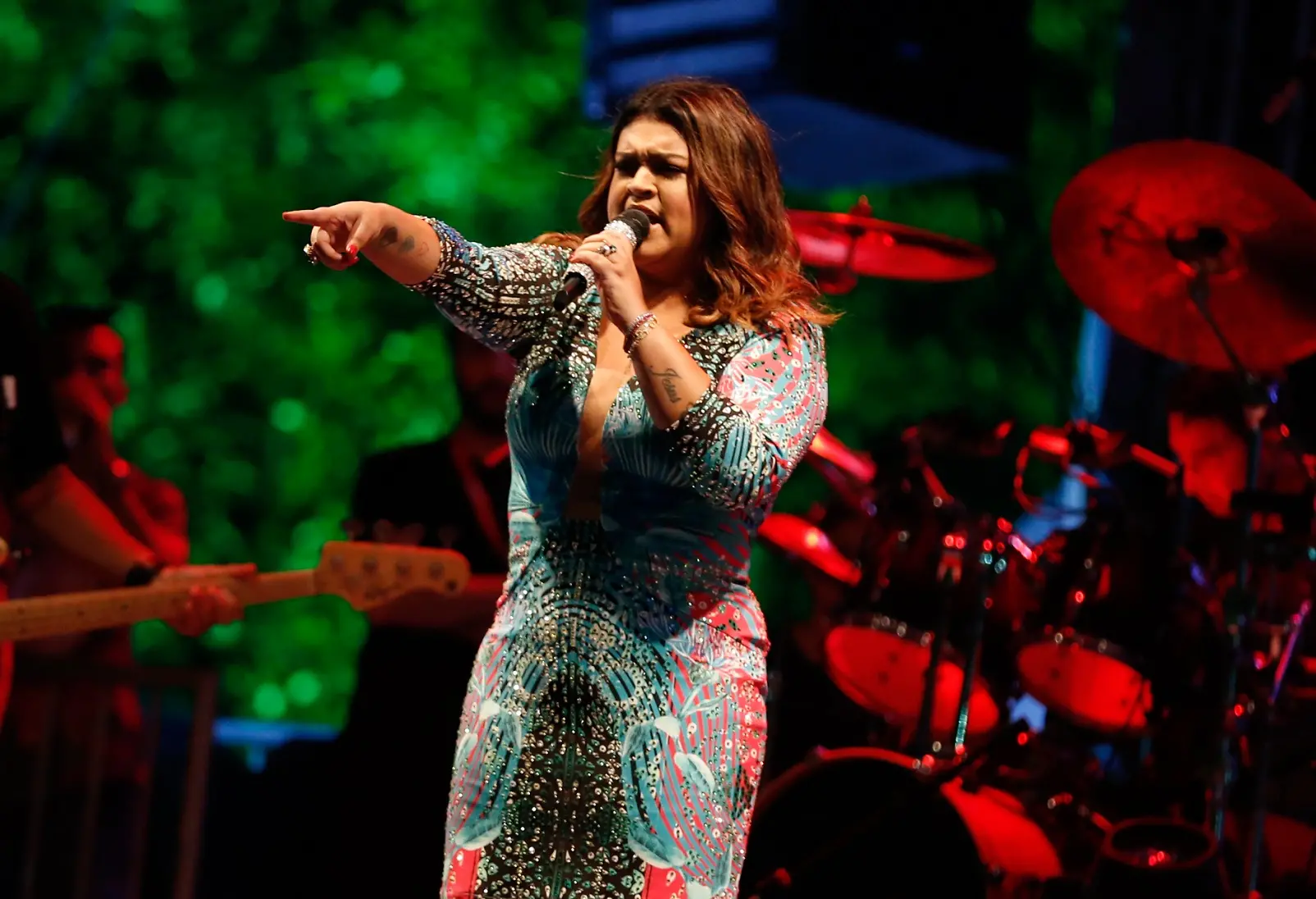 Foto da cantora Preta Gil cantando e segurando microfone em palco. Ela usa um vestido colorido com decote colado ao corpo e aponta o dedo indicador para o público. Atrás dela, uma luz vermelha ilumina a banda