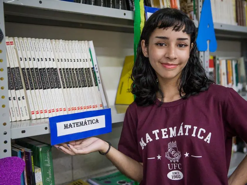 Estudante Rebeca Freitas de Castro, em biblioteca, ao lado de prateleira de livros de matemática