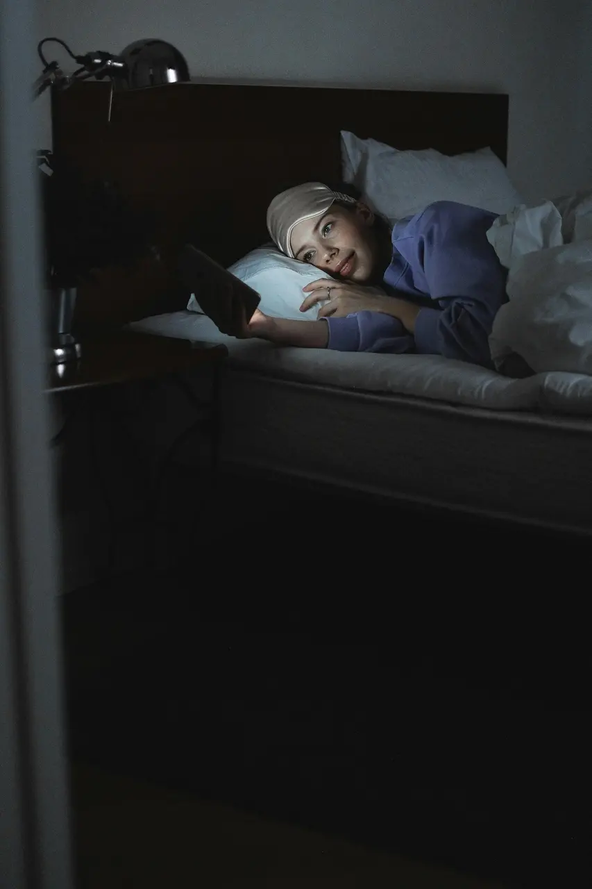 Pessoa deitada na cama em um quarto escuro, usando máscara de dormir na testa. Ela segura um celular iluminado, olhando para a tela com atenção. A luz do aparelho é a principal fonte de claridade na cena.