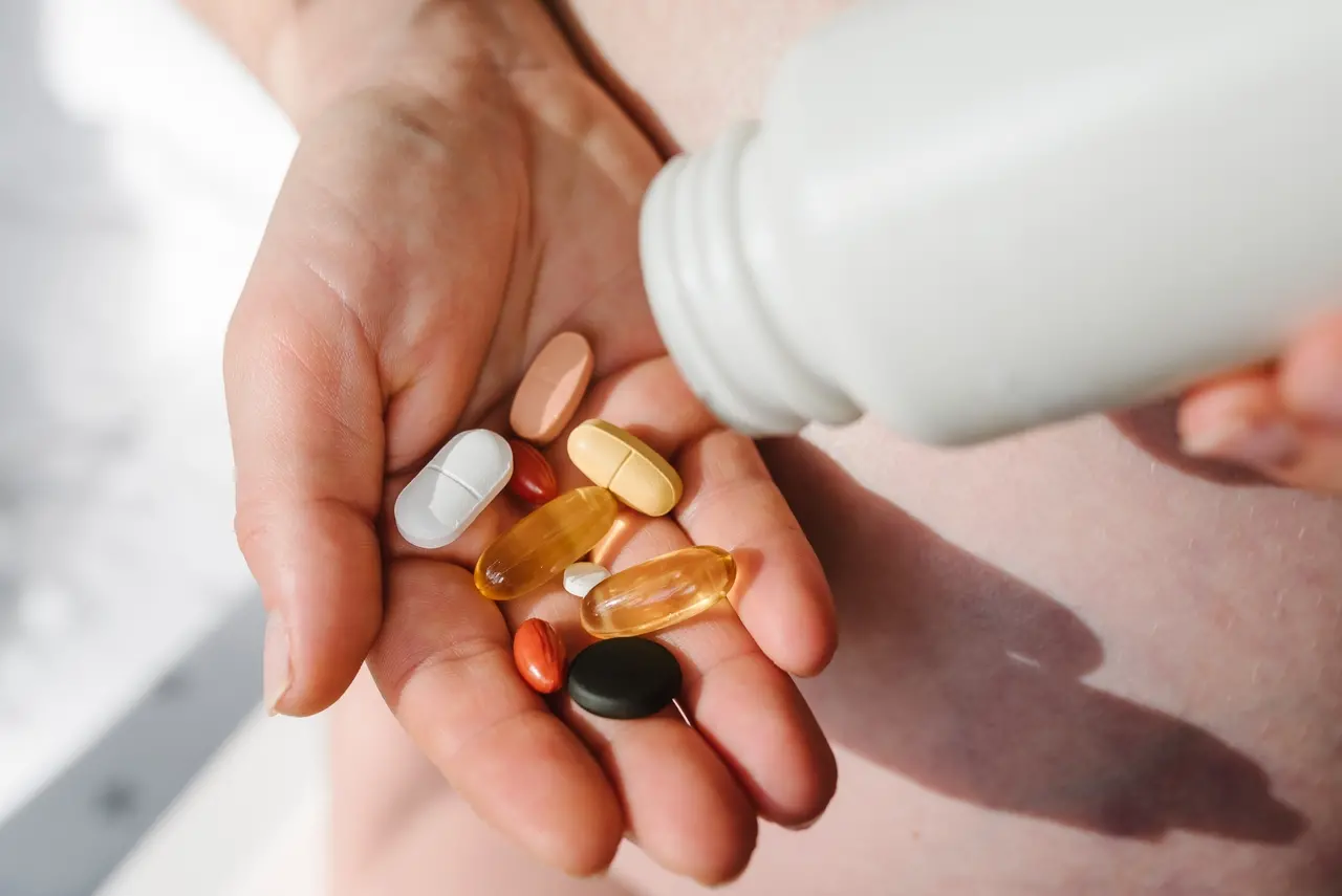 Imagem de uma mão segurando diversos tipos de suplementos vitamínicos e medicamentos, enquanto uma pessoa despeja uma cápsula de um frasco branco, ilustra decisão da ANVISA em proibir venda de sumplentos de uma marca