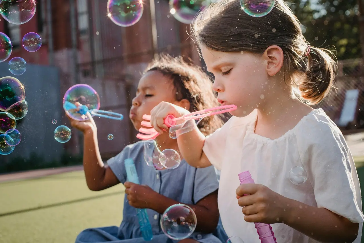 Duas meninas brincam com bolhas de sabão sob a luz do sol. A menina em primeiro plano, com cabelos claros, sopra bolhas de um canudo rosa. A outra menina, em segundo plano, com cabelos escuros, também sopra bolhas.