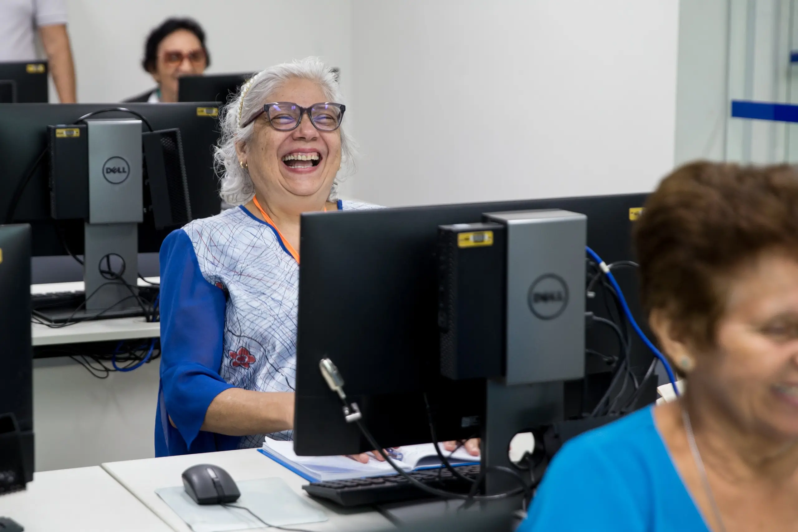 Imagem mostra em plano mais fechado uma sala com idosos sentados na frente de computadores. Esta imagem foca em uma idosa de cabelos brancos e óculos sorrindo.