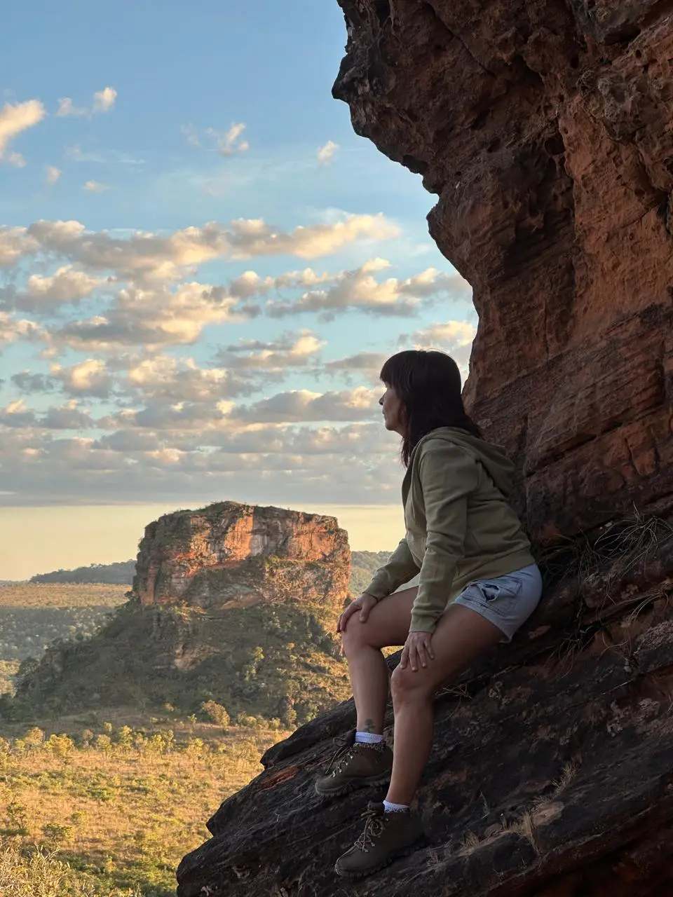Mulher sentada em uma formação rochosa enquanto admira o horizonte. Céu com nuvens e uma montanha está à esquerda da foto.