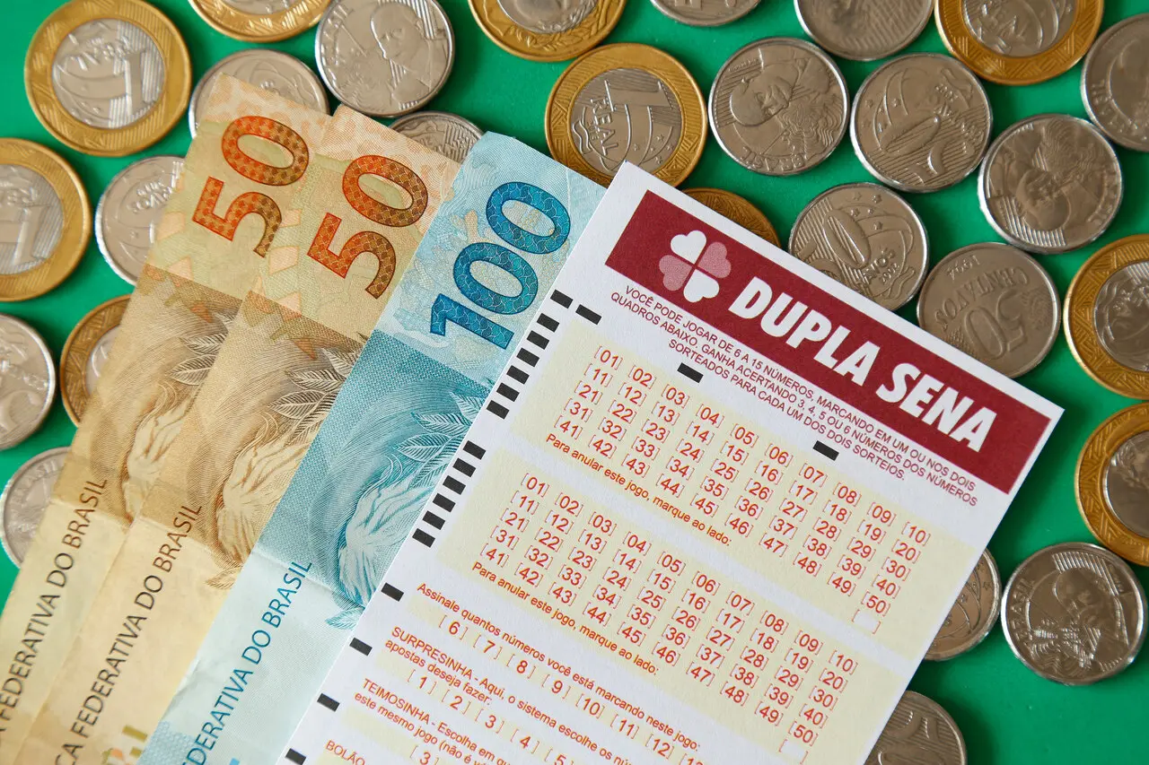 Imagem de uma aposta na loteria Dupla Sena, com dinheiro em moedas e notas abaixo.