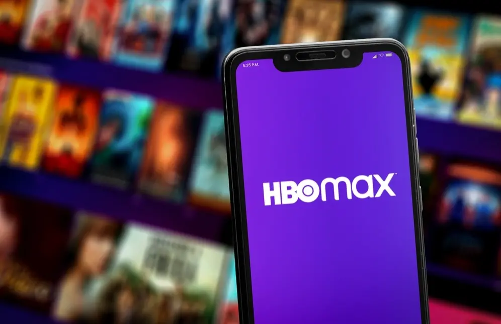 Aplicativo da HBO Max em celular na frente de uma televisão com diversos cartazes de filmes.