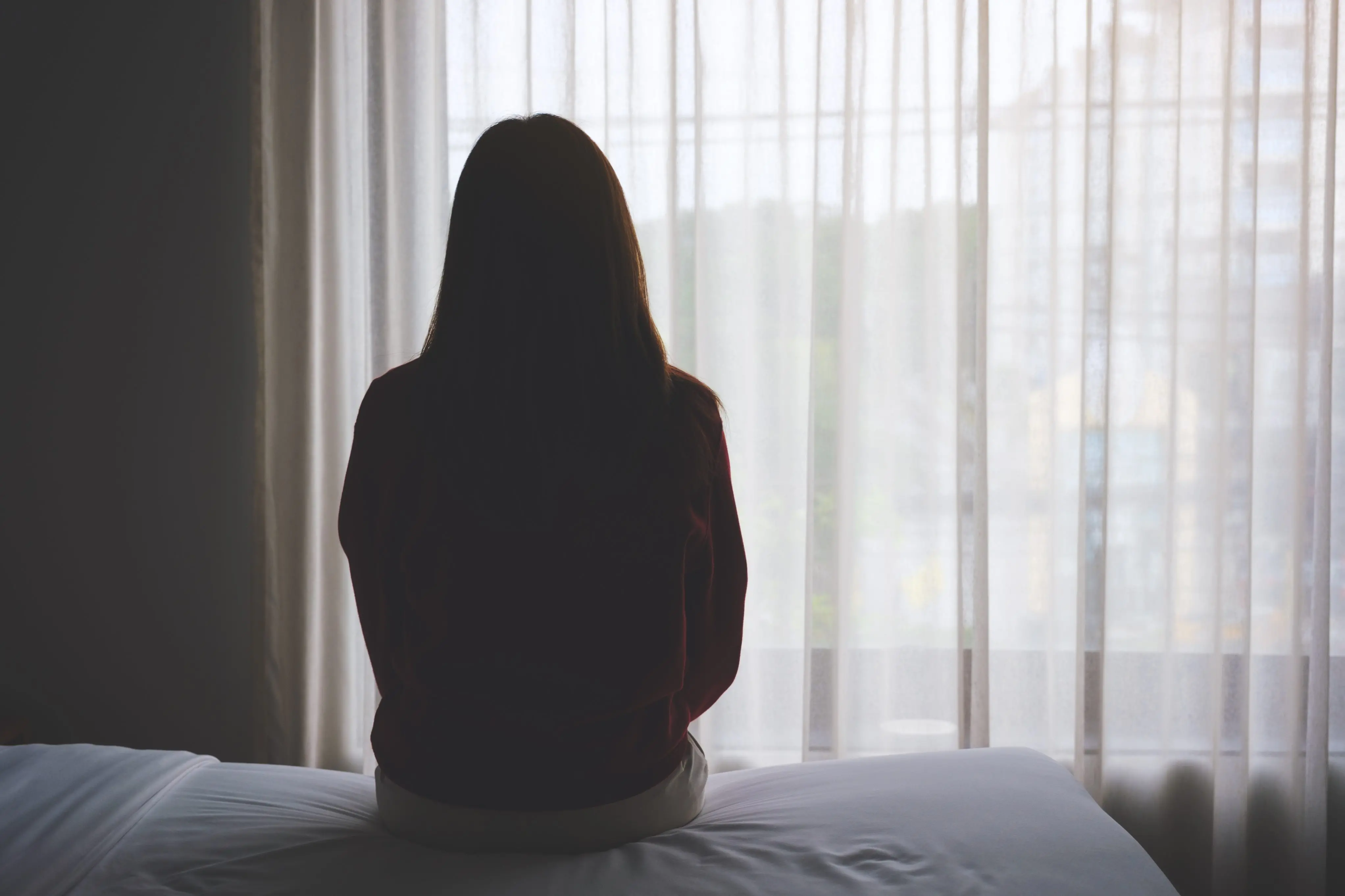Silhueta de uma mulher sentada em uma cama, de costas, olhando para uma janela coberta por cortinas transparentes em um ambiente escuro.