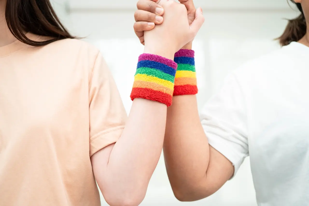 Imagem destaca duas pulseiras com as cores do arco-íris, simbolo da bandeira LGBTQIAP+, no braço de duas pessoas. Não aprarece rossto na imagem.
