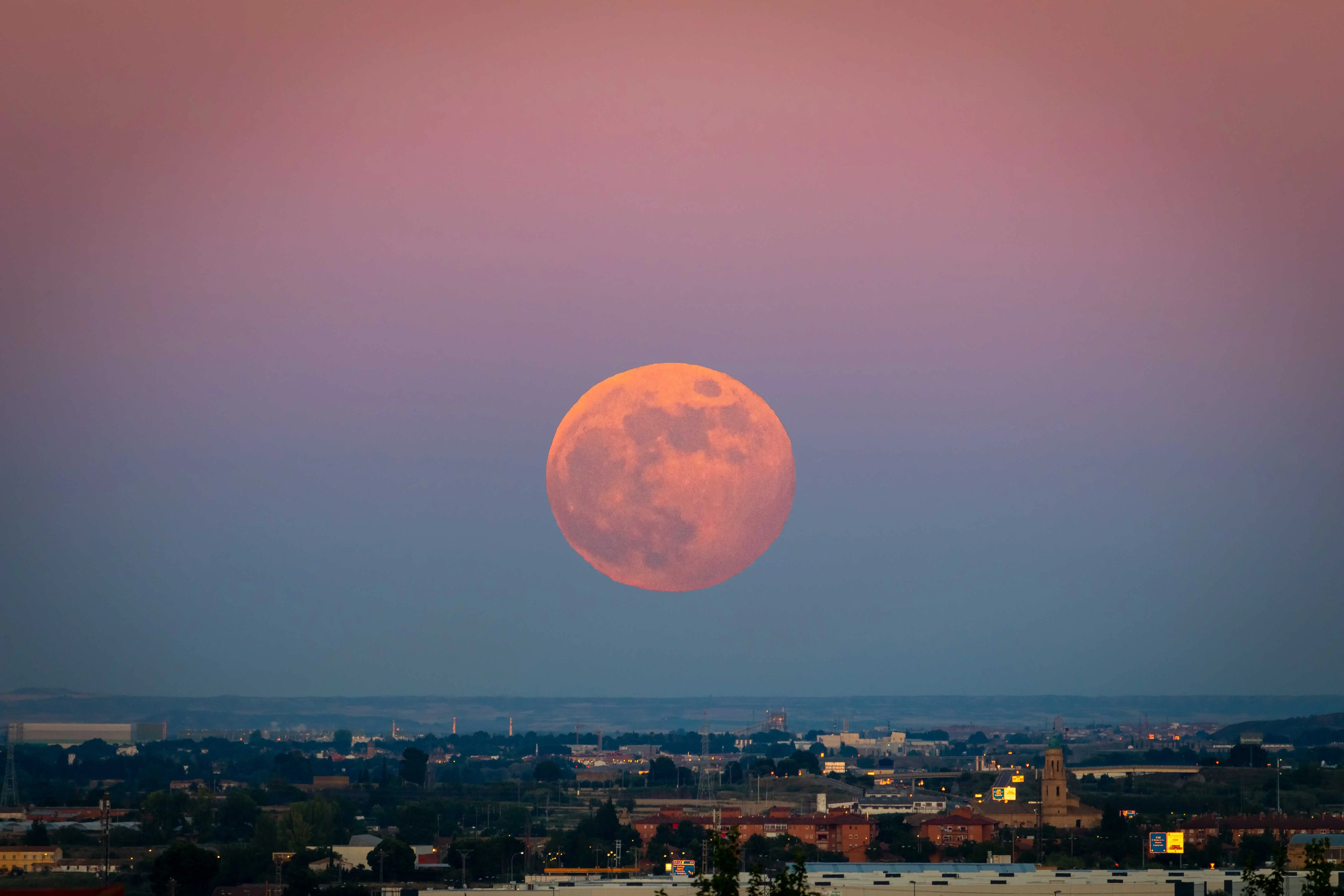 Imagem mostra superlua cheia grande e alaranjada no céu gradiente roxo e azul, pairando sobre o horizonte de uma cidade.