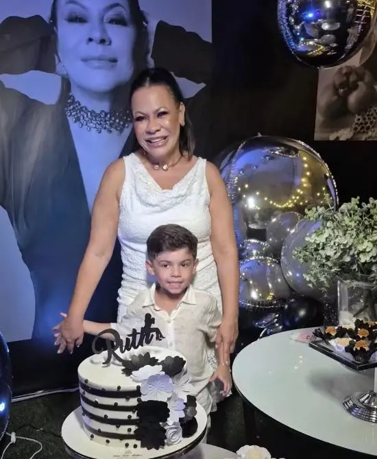 Foto mostra dona ruth e o neto, Léo, sorrindo diante de bolo de aniversário. o bolo tem, no topo, o nome ruth. os dois vestem roupa branca.