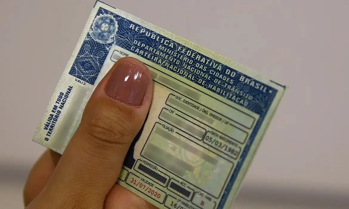 Imagem mostra mão feminina segurando uma Carteira Nacional de Habilitação (CNH) brasileira, com informações pessoais borradas.