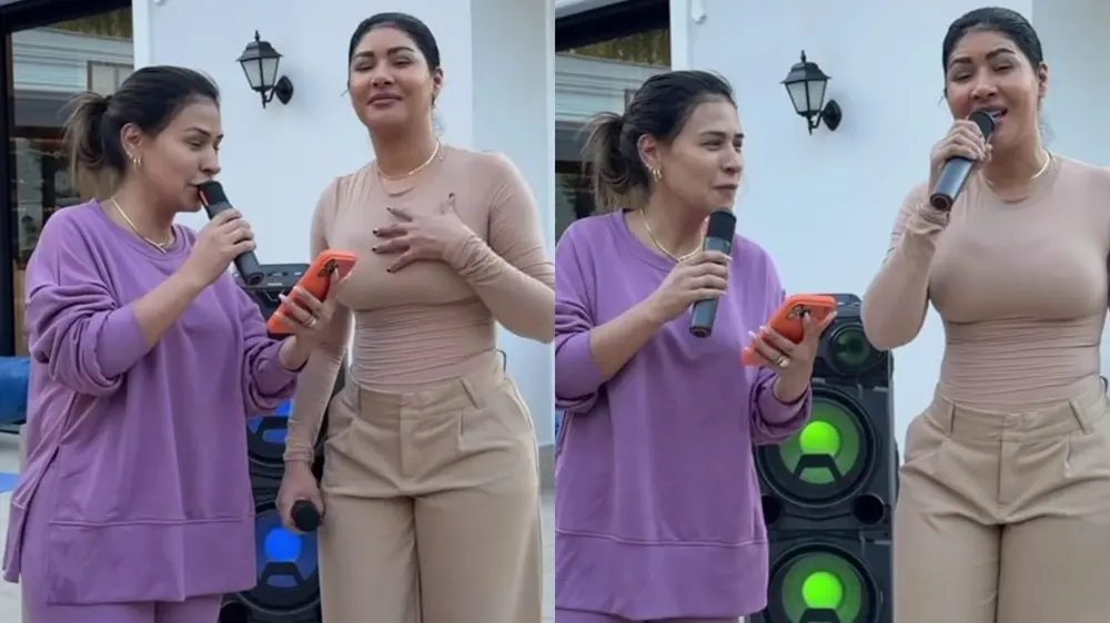 Na imagem, um close-up de duas mulheres, Simone e Simaria, cantando juntas em um evento ao ar livre. A mulher à esquerda (Simaria) usa um conjunto de moletom roxo, segura um microfone na mão direita e um celular na esquerda. A mulher à direita (Simone) usa uma blusa justa cor de pele e calças cáqui de cintura alta, segura um microfone e gesticula com a mão esquerda sobre o peito. Ambas estão de pé, uma ao lado da outra, em frente a uma caixa de som, parecendo felizes e envolvidas na performance. A foto é dividida em duas colunas verticais, com as cantoras posicionadas de forma ligeiramente diferente em cada uma, sugerindo que é um momento de um vídeo.