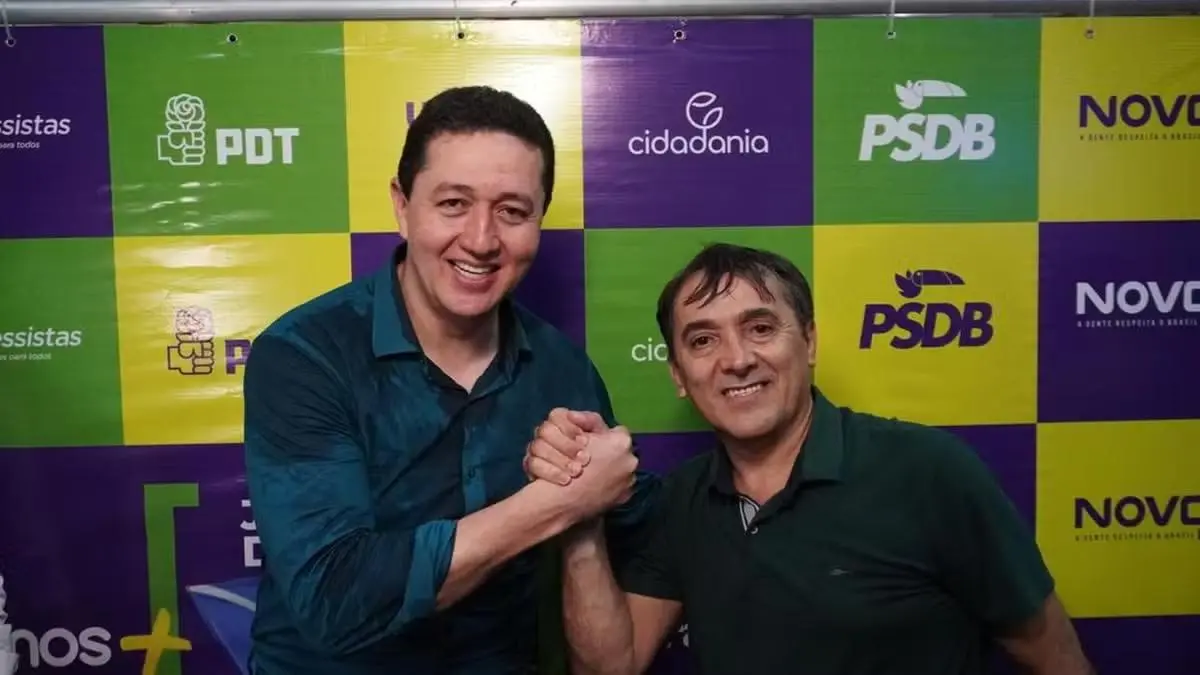 Dois homens sorrindo e apertando as mãos em gesto de cumprimento diante de um painel colorido com logos de partidos políticos, incluindo PDT, Cidadania, PSDB e Novo.