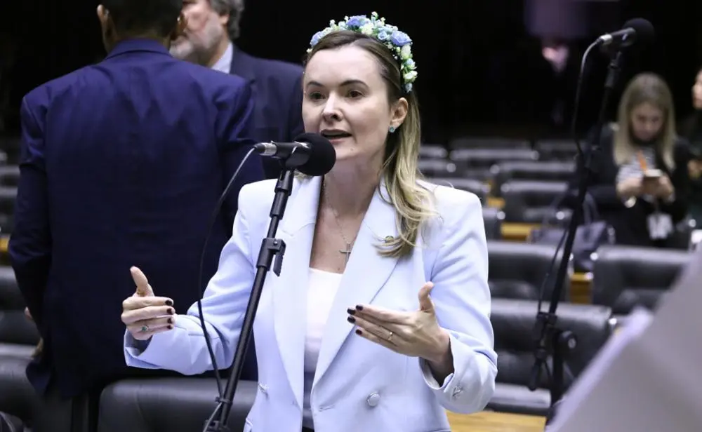 A deputada federal Júlia Zanatta fala ao microfone em um ambiente legislativo. Ela veste um blazer azul-claro e usa uma coroa de flores na cabeça, gesticulando com as mãos enquanto se dirige ao plenário. Outras pessoas aparecem desfocadas ao fundo.