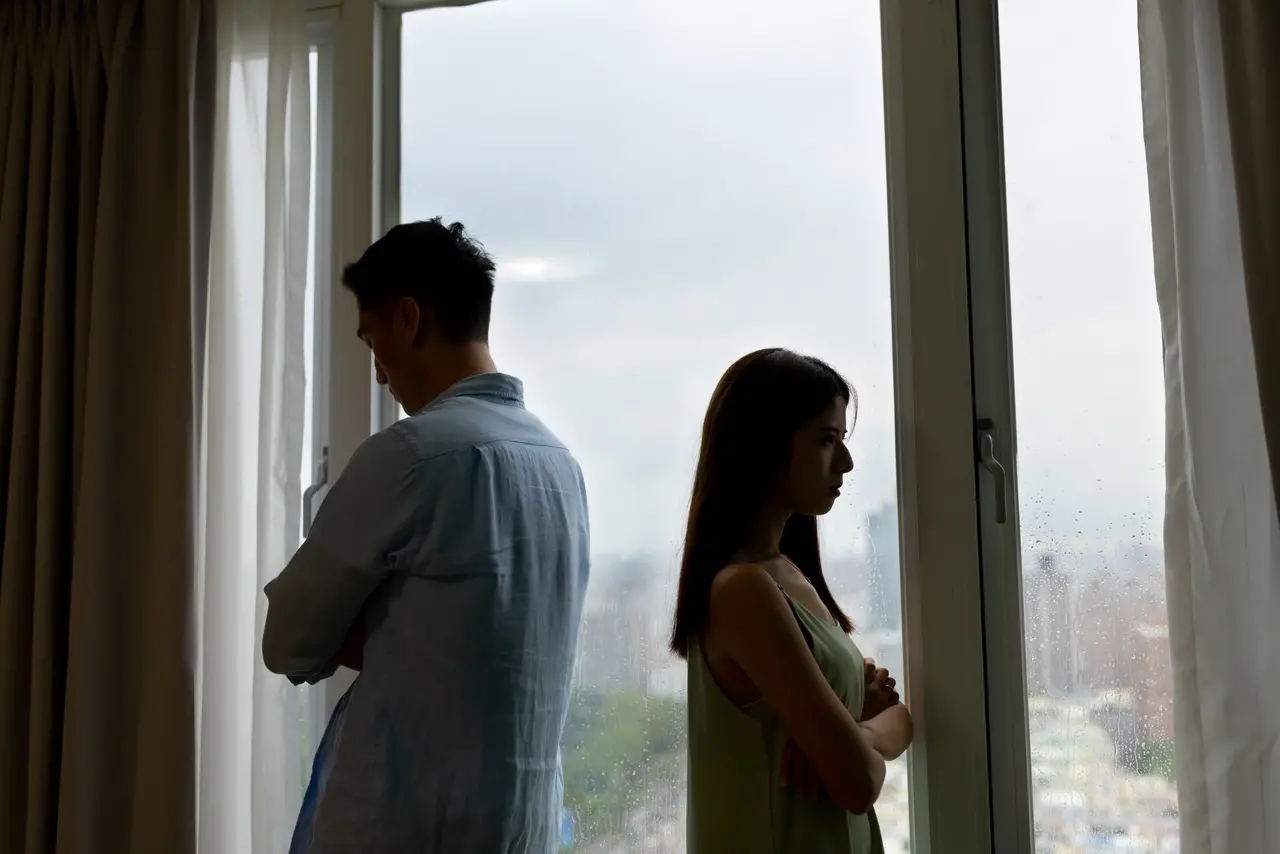 Foto de casal formado por um homem e uma mulher com destaque para as silhuetas dos dois, que estão distantes em frente a uma janela de vidro.