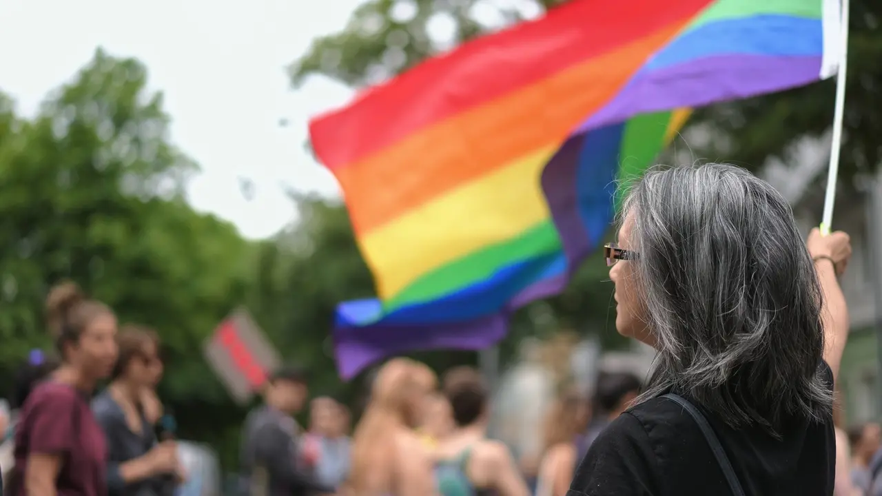 Mulher idosa com cabelos grisalhos, óculos e vestindo preto, vista de costas e de perfil, em uma parada da diversidade. Ela segura uma grande bandeira do orgulho arco-íris (LGBT) que flutua no fundo. Ao seu redor, há uma multidão de pessoas borradas, em um ambiente exterior com árvores.