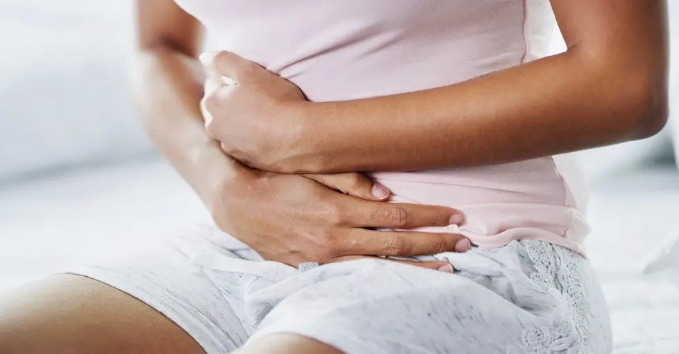Mulher negra com dor segura a barriga. Ela está de short cinza e camiseta rosa.