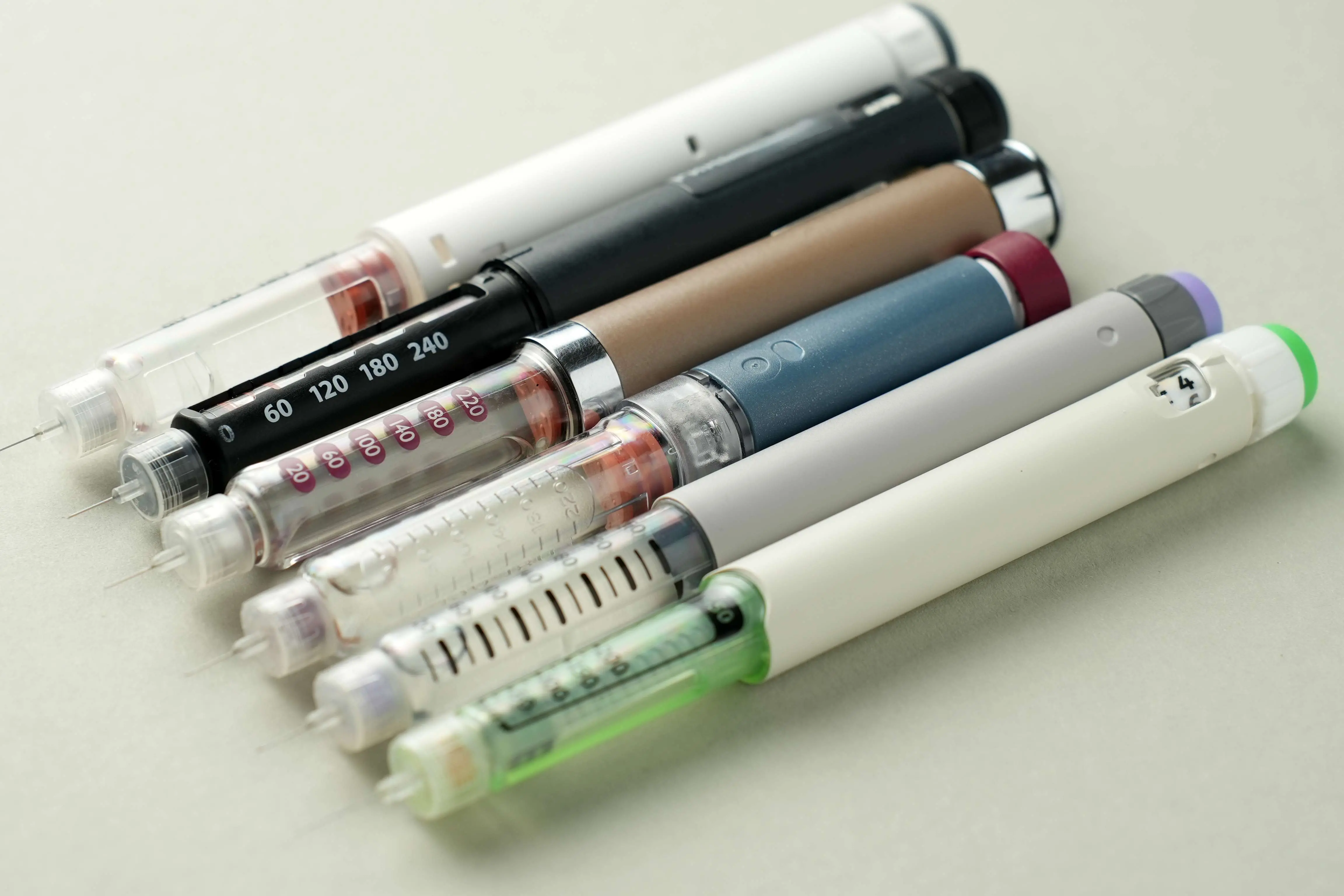 Imagem mostra diversos tipos de agonistas da GLP-1, conhecidos como canetas emagrecedoras, de diferentes cores e tamanhos em uma superfície branca, usados para administração de medicamentos.