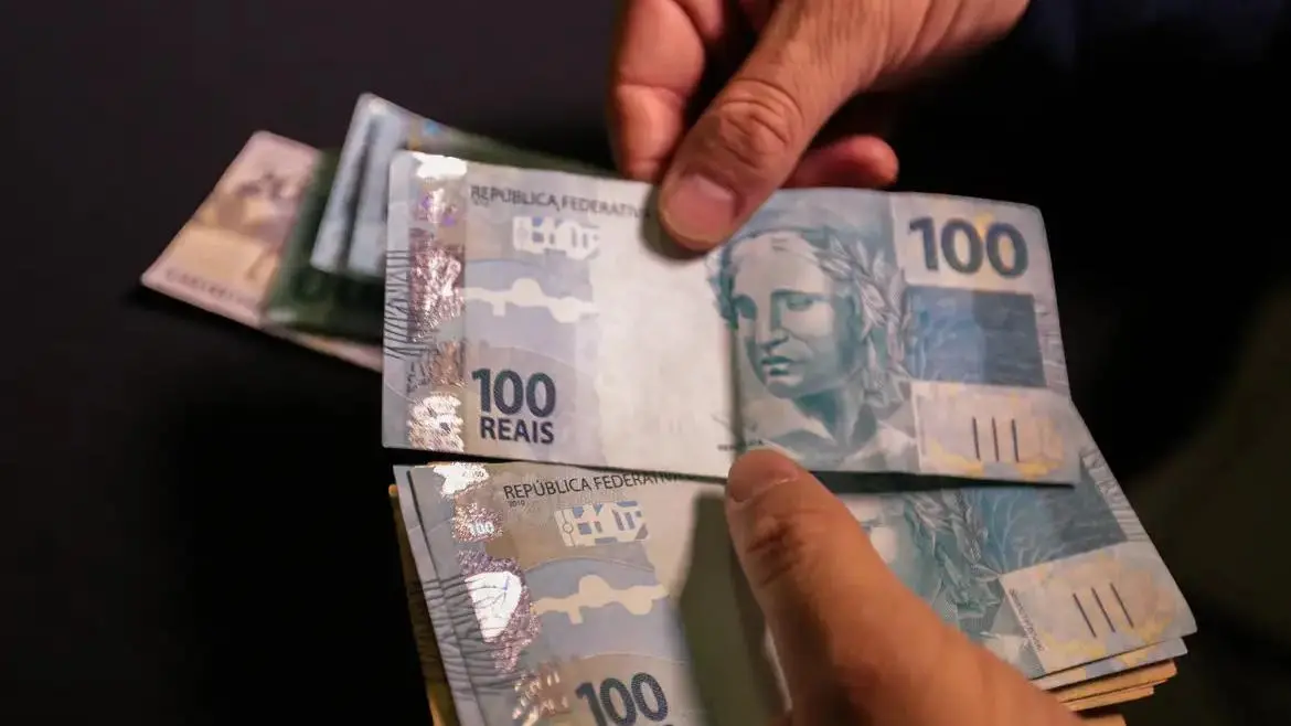 Imagem mostra uma mão que segura cédulas de R$ 100.