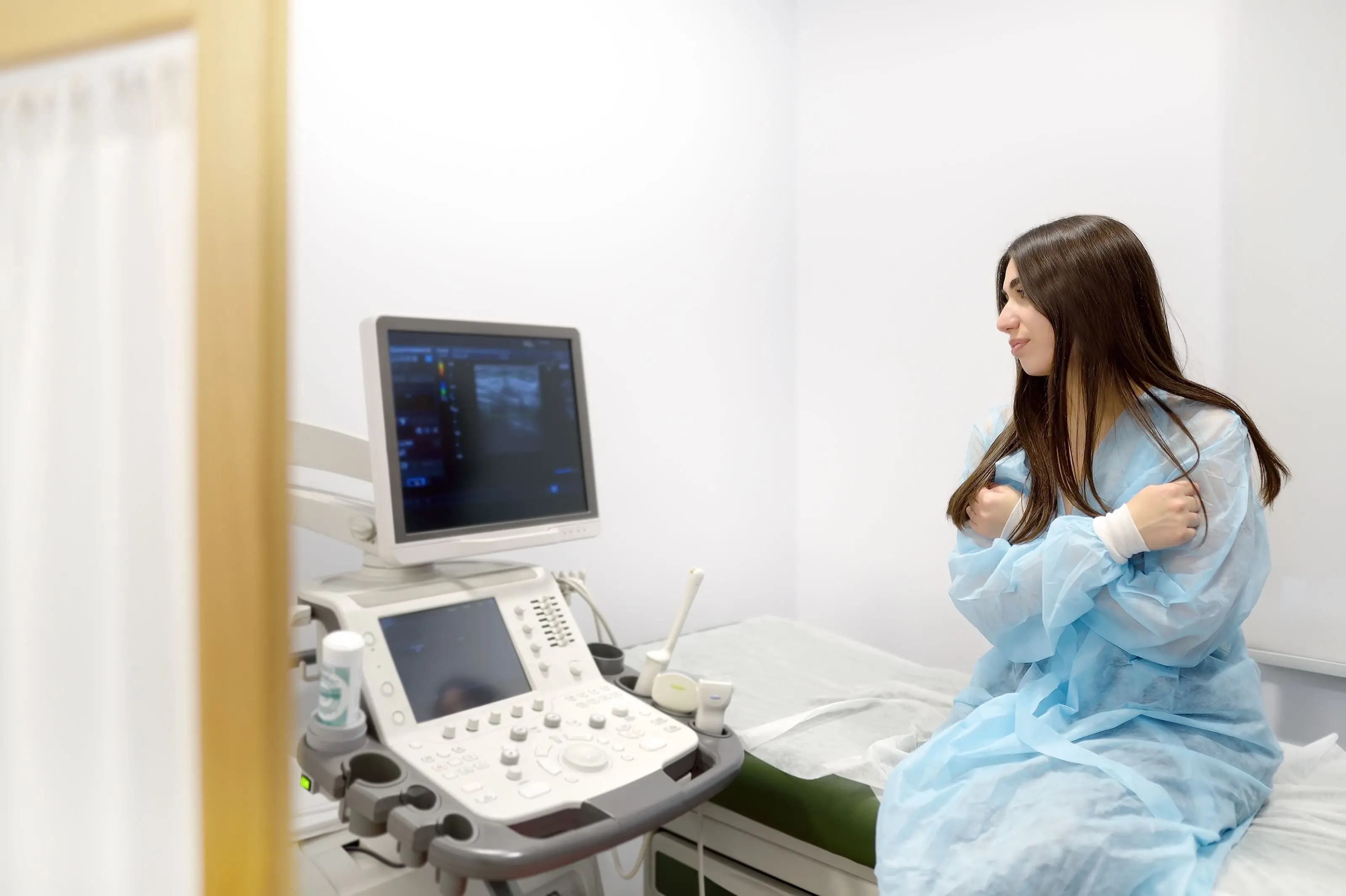 Mulher jovem em avental hospitalar sentada em maca, ao lado de um aparelho de ultrassom em um consultório médico.