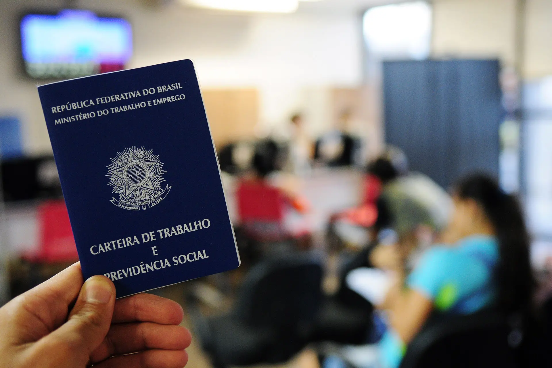 Primeiro plano de uma Carteira de Trabalho brasileira sendo exibida, simbolizando emprego e direitos trabalhistas, com fundo de público em escritório.