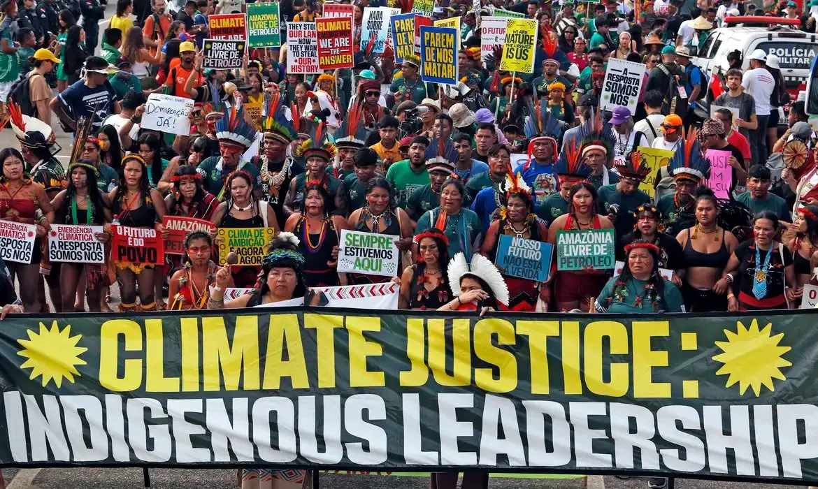 Multidão em manifestação por justiça climática e liderança indígena, com uma grande faixa em primeiro plano e participantes diversos, durante a COP30 em Belém, no Pará.