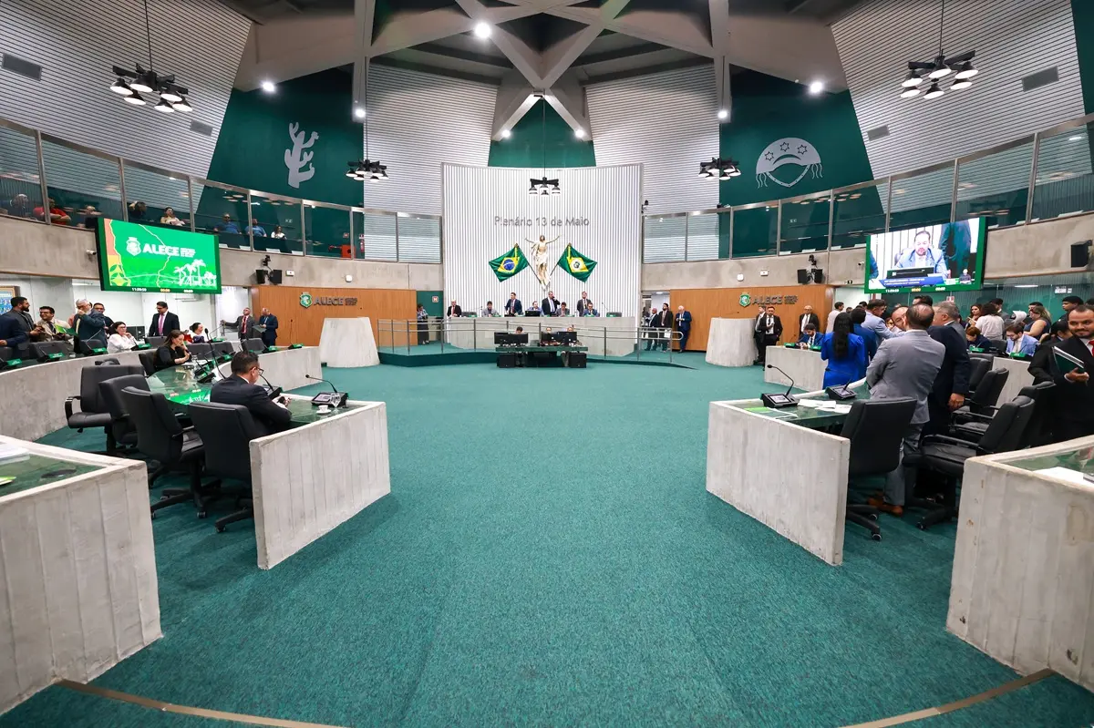 Plenário da Assembleia Legislativa do Ceará durante sessão, com parlamentares distribuídos em bancadas semicirculares. Ao fundo, mesa diretora diante de duas bandeiras do Brasil e do Ceará. Telões laterais exibem a transmissão da sessão. Pessoas conversam em grupos no lado direito do plenário.